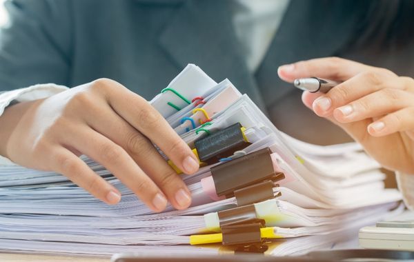 A stack of paperwork held together in individual documents by coloured paperclips or bull dog clips. You can see the hands of someone reaching for the paperwork and holding a pen