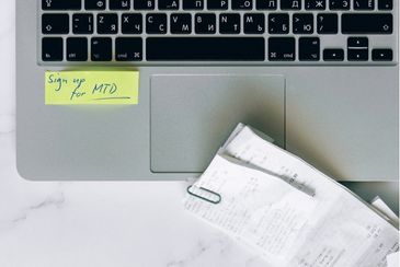 A laptop keyboard with a post it note that says sign up for MTD written on it in blue ink and a small bundle of till receipts held together with a paper clip